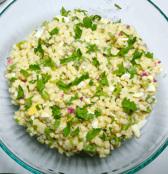 A clear bowl set on top of a white board filled with pearl couscous mixed with cuts of asparagus, chopped cooked egg white, pops of diced red onion, all tossed in lemon-parmesan vinaigrette, and garnished with bright green chopped parsley. A delicious side salad for a picnic lunch date.