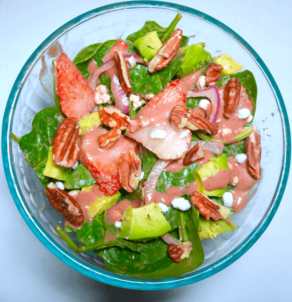 Vibrant, green spinach leaves mixed with a dark pink with a hint of brown strawberry balsamic vinagrette, topped with three slices of strawberry, chopped pecans, chunks of creamy avocado, and goat cheese crumbles. A few slices of red onion are apparent on top, and the entire salad is drizzled with the vinagrette. This salad is displayed on a white surface inside of a clear salad bowl. 